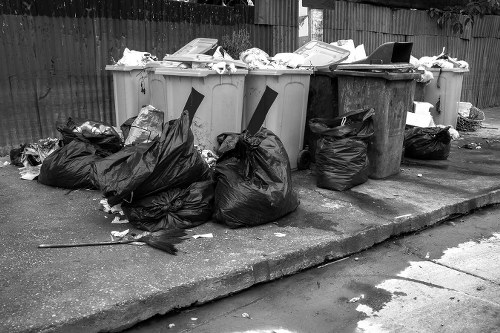 A group of large black and grey rubbish bags filled with waste materials sit on the pavement next to a row of vehicle-sized wheelie bins, including grey and black bins with open lids, in an outdoor area adjacent to a wooden fence. The bags appear to contain discarded household or commercial waste and are placed on a concrete surface with some debris scattered around. Behind the bins, the wooden fence extends across the background, suggesting the waste is in a residential or commercial yard. The scene is captured in black and white, emphasizing the textures of the waste bags, bins, and pavement, with natural daylight illuminating the area. This image illustrates typical rubbish disposal and collection points, relevant to commercial waste removal services like those offered by Commercial Waste Grays in Grays, Essex.