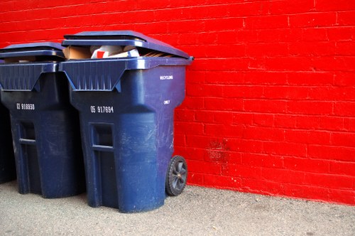 Two large dark blue wheeled rubbish bins made of durable plastic are positioned side by side on a concrete surface next to a red brick wall. The bins feature textured surfaces with vertical ridges and are fitted with black lids, one of which is slightly open, revealing some mixed waste materials inside. The bins are placed in an outdoor area, possibly a back alley or service yard, with minimal surrounding environment visible. The setting suggests a commercial or residential waste collection point, consistent with rubbish removal services offered by Commercial Waste Grays. The lighting is neutral, highlighting the clean lines and slightly glossy finish of the bins, emphasizing their utilitarian purpose in waste management. The image captures the practical aspect of rubbish disposal, aligning with the company's service category of rubbish removal in Grays, Essex, providing a clear visual reference for customers seeking waste disposal solutions in the local area.