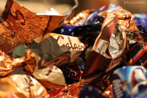 A close-up image showing a collection of crumpled foil and plastic wrappers, primarily in metallic copper, silver, and various vibrant colours, piled together. The wrappers display different textures with shiny, reflective surfaces and some printed text, suggesting they are confectionery or snack wrappers. The background is blurred, emphasizing the foreground clutter of packaging materials. This collection of discarded wrappers is typical of waste generated from food items and treats, relevant to rubbish removal services like those offered by Commercial Waste Grays operating in Grays. The environment appears to be indoors, possibly on a table or in a waste collection context, with the focus on the waste material intended for disposal or collection. The detailed textures and mixture of metallic and colourful finishes highlight the types of household or commercial rubbish that require professional rubbish removal, supporting services offered in the Grays area.