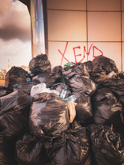 A person wearing a long-sleeved, light-colored striped shirt is holding a large, transparent plastic bag filled with various clear and coloured plastic bottles and containers. The bag is gripping the 