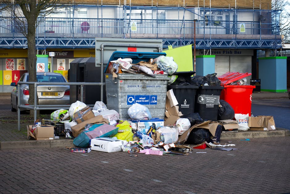 A large pile of black rubbish bags, some torn open exposing contents such as crumpled paper and plastic bottles, is stacked against a contemporary building with a sleek, tiled exterior in an urban set