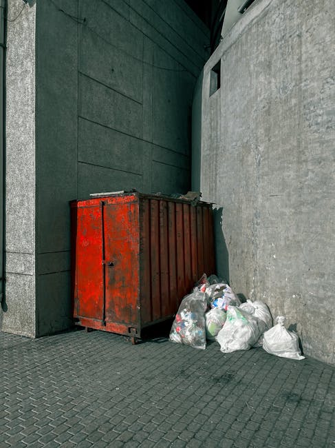 The image depicts a red, weathered metal waste container placed against a dark green building wall with a rough texture, situated on a paved sidewalk. To the right of the container, several white plas